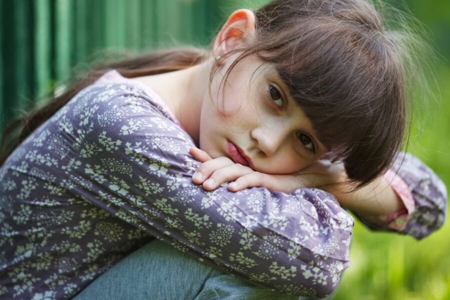 A sullen girl rests her head on her knees while sitting outdoors.