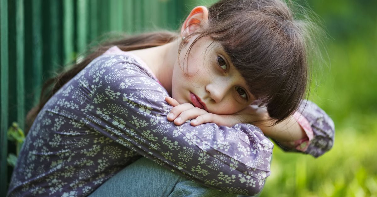 A sullen girl rests her head on her knees while sitting outdoors.
