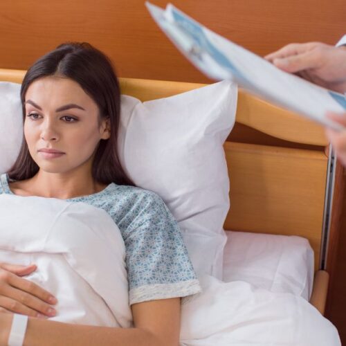 A woman lays in a hospital bed while a doctor talks to her.