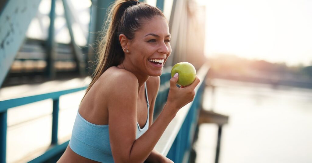 A woman in exercise clothing eats an apple while on a bridge looking out over a river.