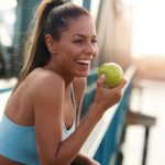 A woman in exercise clothing eats an apple while on a bridge looking out over a river.