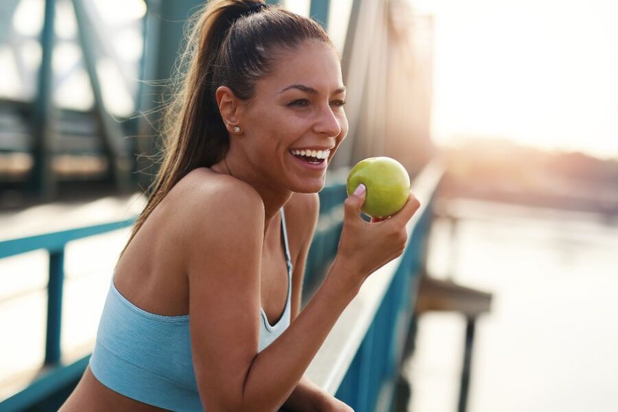 A woman in exercise clothing eats an apple while on a bridge looking out over a river.