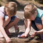 Two girls play in the dirt near a stream.