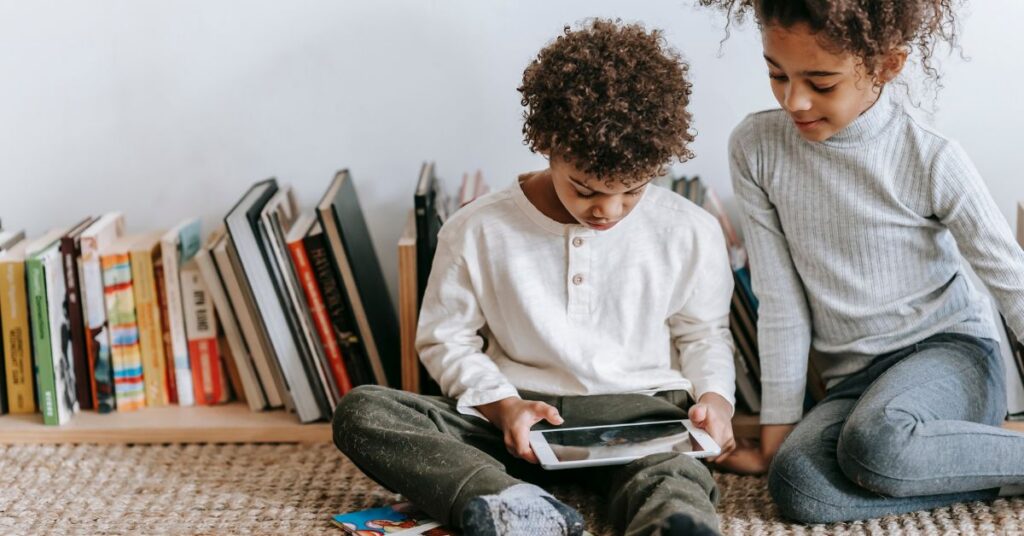 A brother and sister sit on the floor while looking at a tablet.