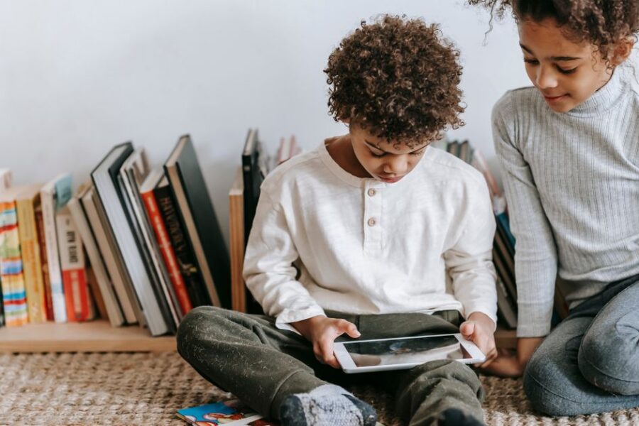 A brother and sister sit on the floor while looking at a tablet.