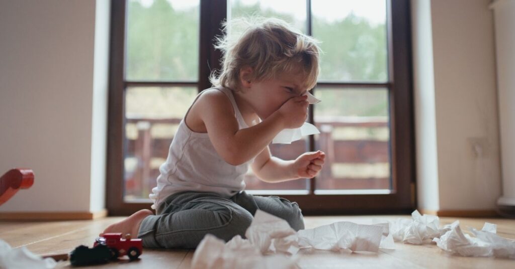 A toddler sitting on the floor surrounded by tissues wipes his stuffy nose.