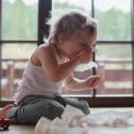 A toddler sitting on the floor surrounded by tissues wipes his stuffy nose.