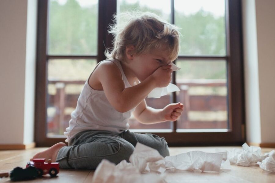 A toddler sitting on the floor surrounded by tissues wipes his stuffy nose.